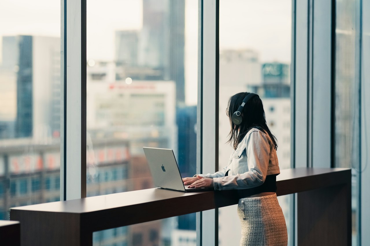 Una persona con cascos trabaja de pie con un portátil junto a un ventanal, con la ciudad desenfocada al fondo.
